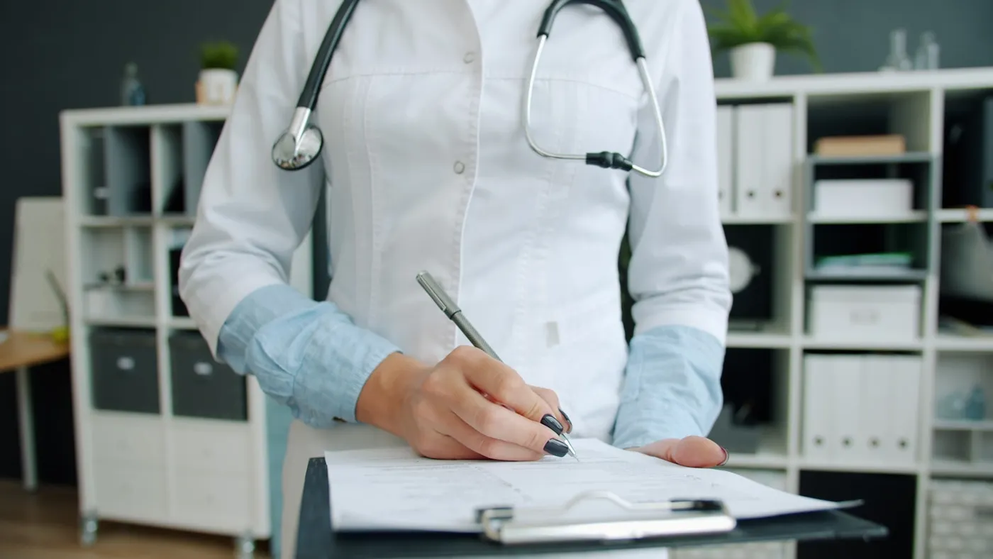 A clinician in a white coat signing paperwork on a clipboard in a private consulting room